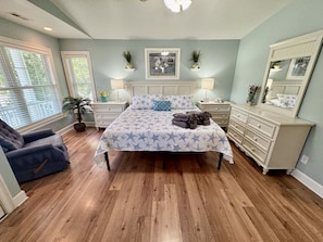 A bedroom with a bed, bedside tables, a dresser, a chair, and a large window. - The Salty Rooster | Ocean Lakes 4BR | Golf Cart + Fire Pit | Sleeps 8 (Myrtle Beach)