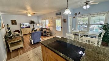 A kitchen with a central island, a dining area with white chairs, and a living room with a sofa and a television.