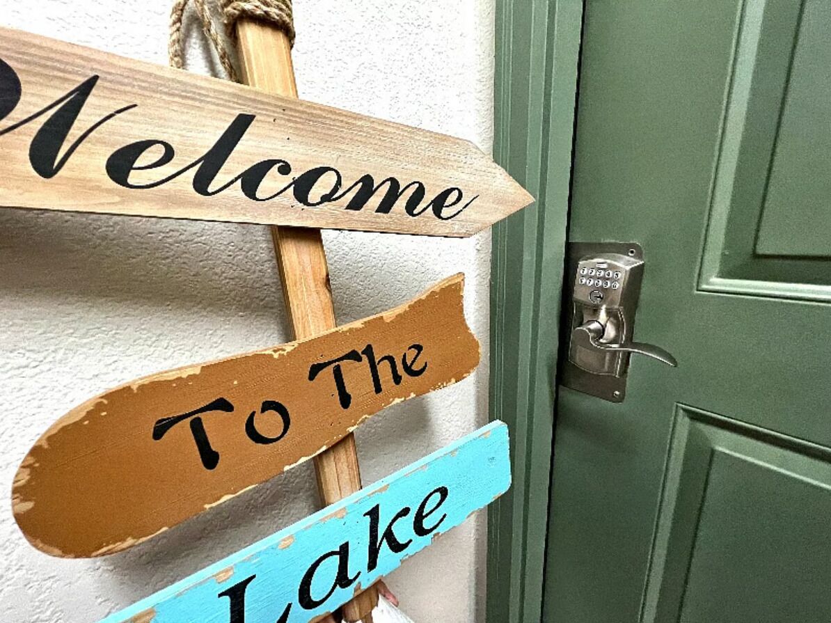 Et træskilt med teksten 'Welcome' og 'To The Lake', monteret på en grøn dør.