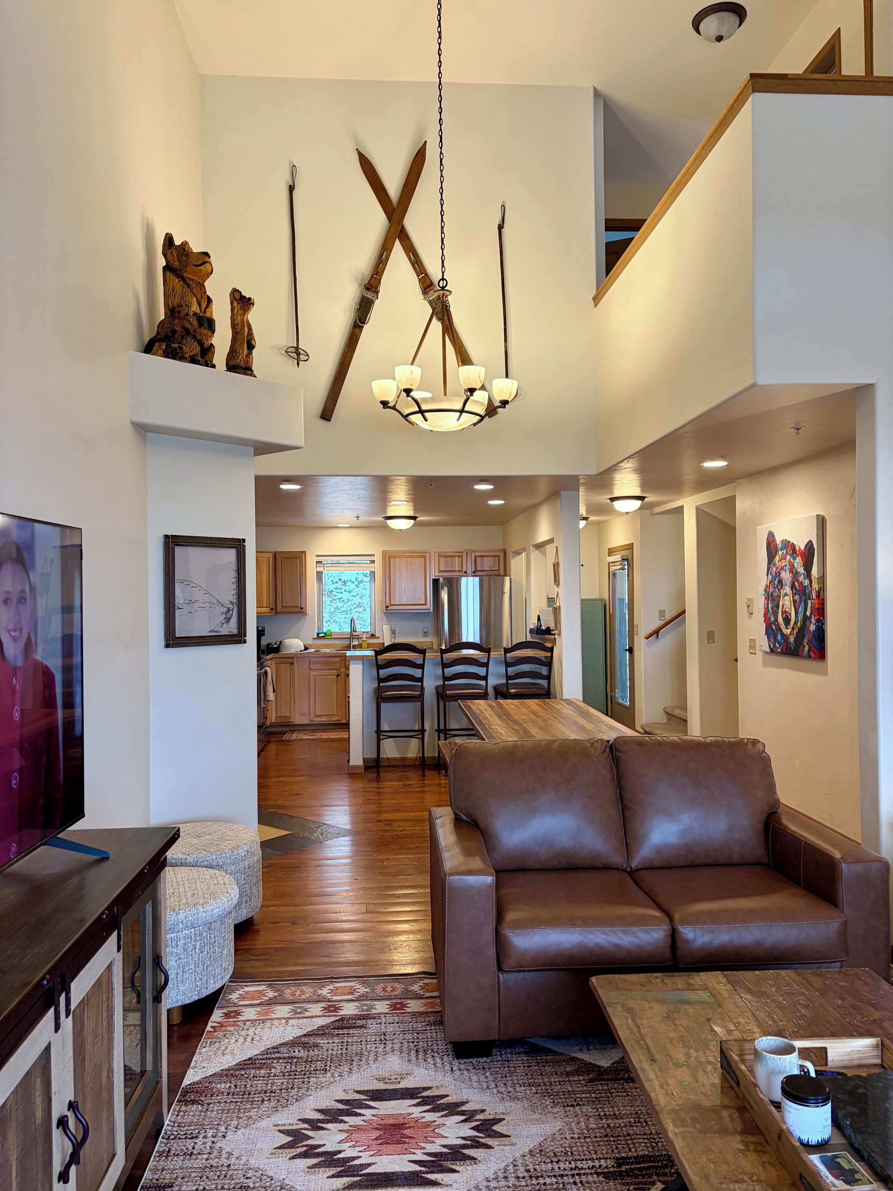 A spacious living area with a brown leather sofa, a wooden coffee table, and a patterned rug. A television is mounted on the wall, and there is a kitchen visible in the background.