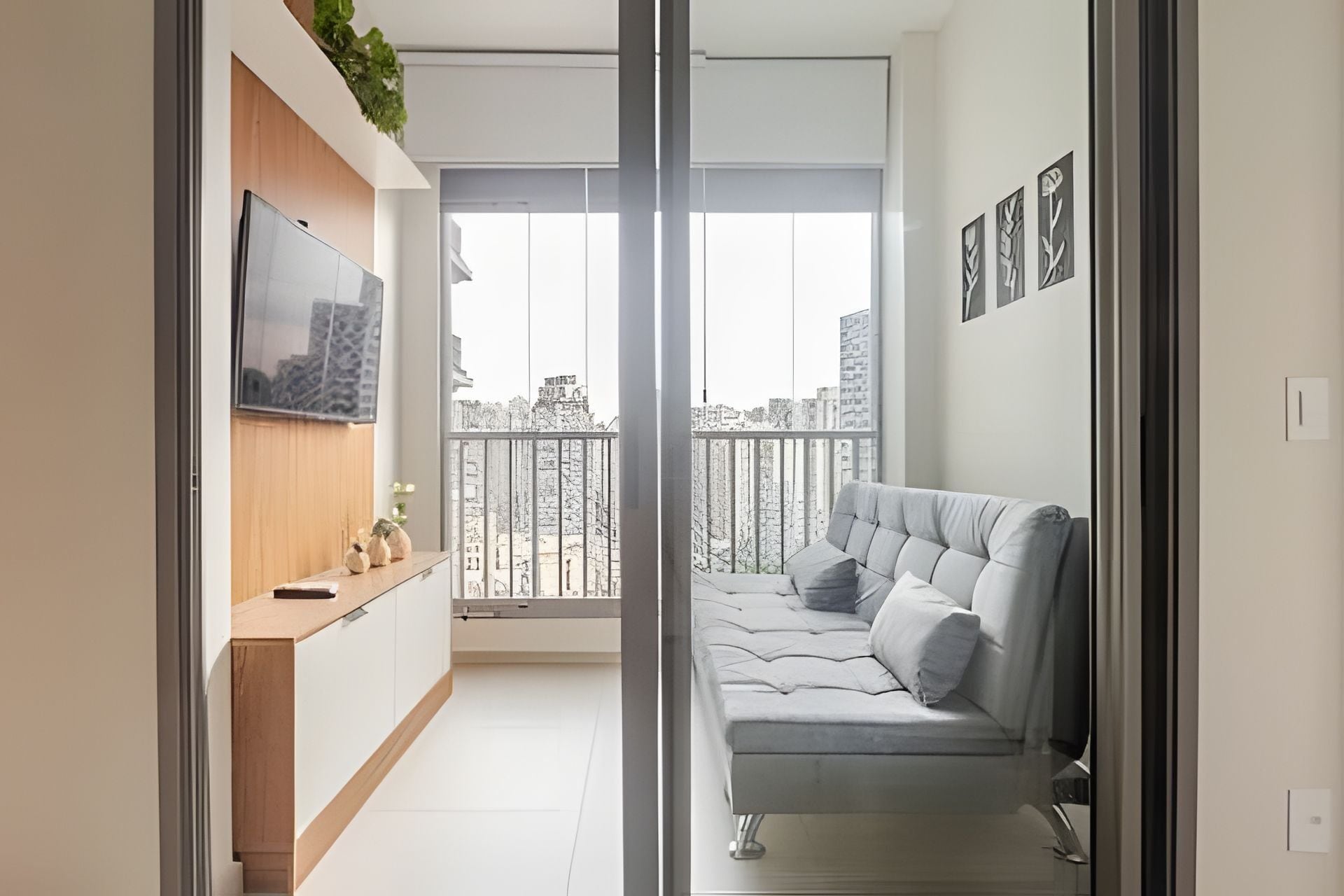 A modern living room with a grey sofa, a flat-screen TV mounted on a wooden cabinet, and a balcony with a view of the city.