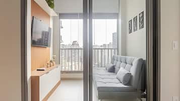 Uma sala de estar moderna com um sofá cinzento, uma televisão de tela plana fixada em um móvel de madeira, e uma varanda com vista para a cidade.
