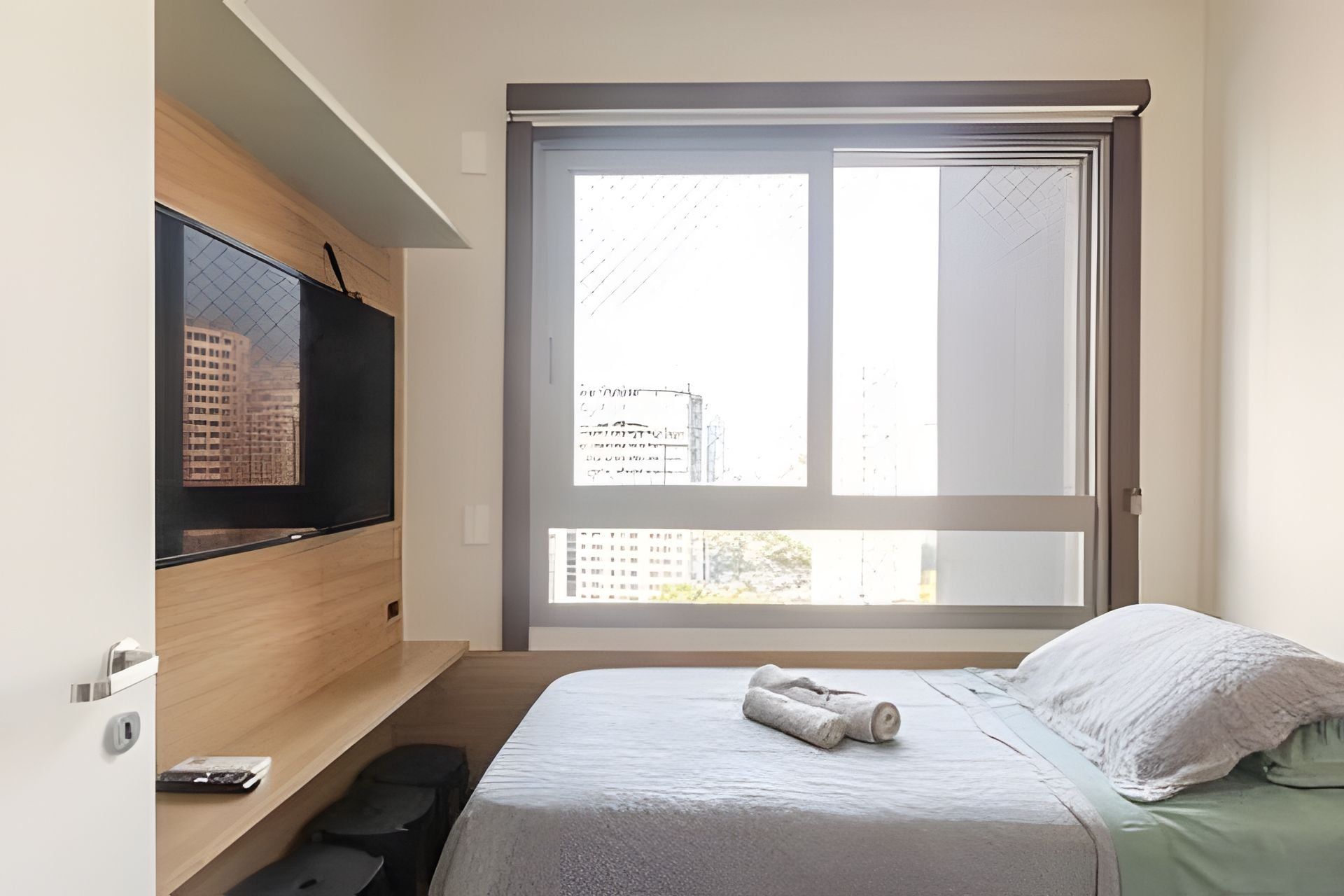 A bedroom with a bed, a TV on a wooden shelf, a large window with a view of buildings, and a towel on the bed.