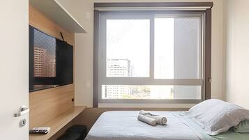 Um quarto com cama, uma TV em uma estante de madeira, uma janela grande com vista para edifícios e uma toalha na cama.