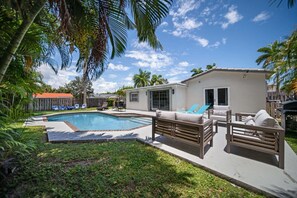 A backyard with a swimming pool, a covered patio with seating, and a small building with windows. - 2600 Managed by Brampton Park (Pompano Beach)