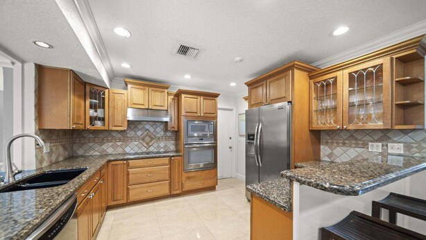 A kitchen with wooden cabinets, granite countertops, stainless steel appliances, and a tiled backsplash. - 2600 Managed by Brampton Park (Pompano Beach)
