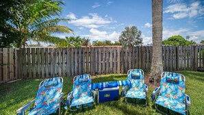 A backyard with a wooden fence, a palm tree, and four lawn chairs with a floral pattern. - 2600 Managed by Brampton Park (Pompano Beach)
