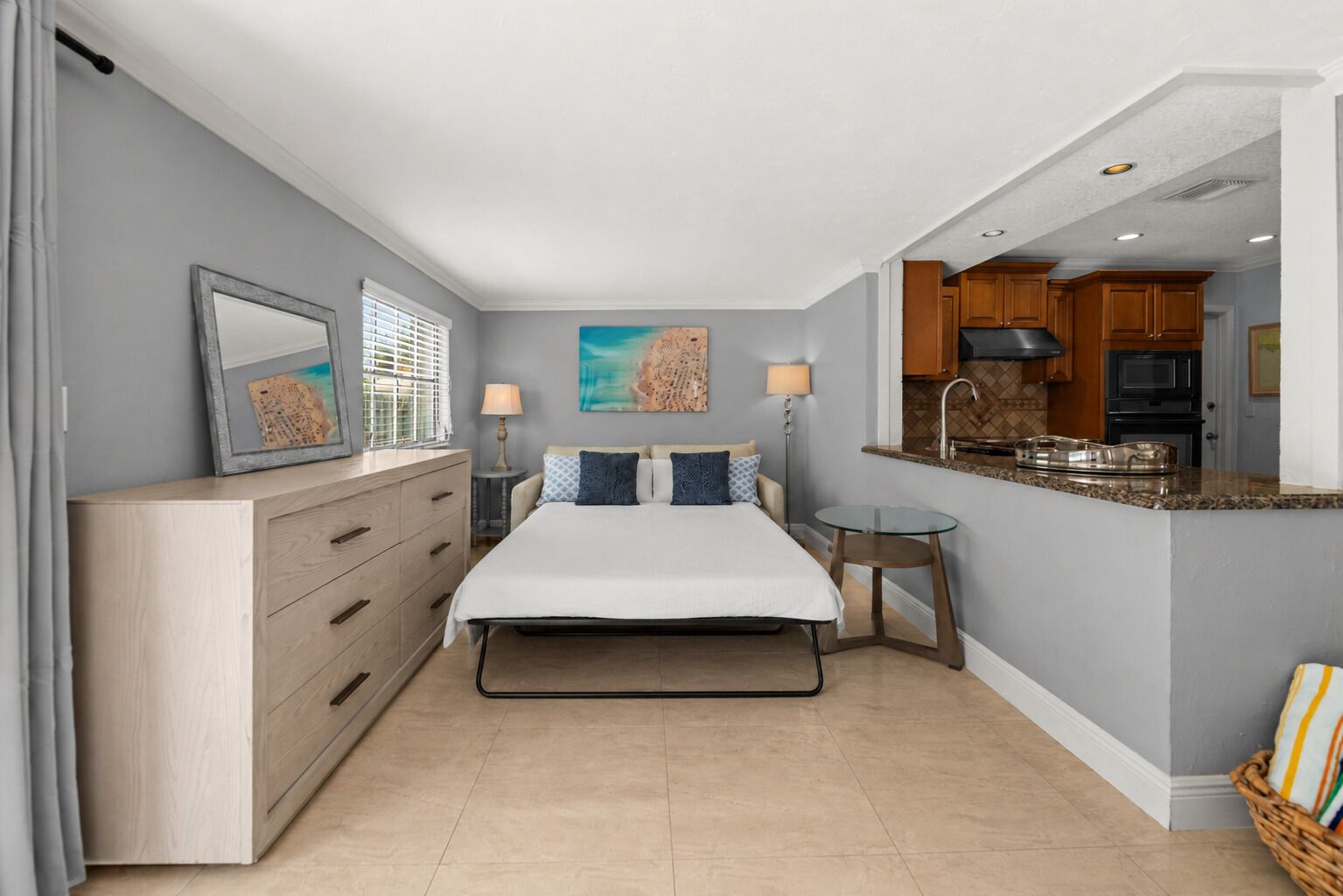 Une chambre à coucher comprenant un lit, des tables de chevet, une commode, une fenêtre avec des stores et une vue sur la cuisine.