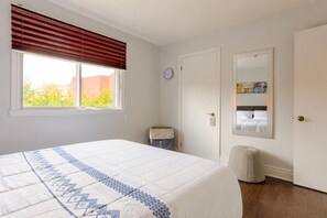 A bedroom with a large bed, a window with blinds, a door with a mirror, and a trash bin. - Cozy 2 bedroom close to downtown, public transportation & airport (Montréal)
