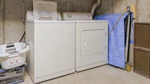 A laundry area with a washing machine, dryer, and a utility room with exposed pipes and unfinished walls. - Cozy 2 bedroom close to downtown, public transportation & airport (Montréal)