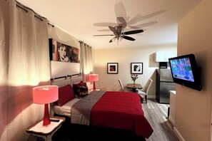 Habitación de hotel con cama, escritorio, silla, televisor, ventilador de techo y refrigerador.