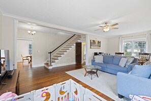 A spacious living room with hardwood flooring, a blue sofa, a glass coffee table, and a ceiling fan. - Historic 1935 Charmin Retreat 4BR Near Devil’s Den and Cedar Lake (Williston)
