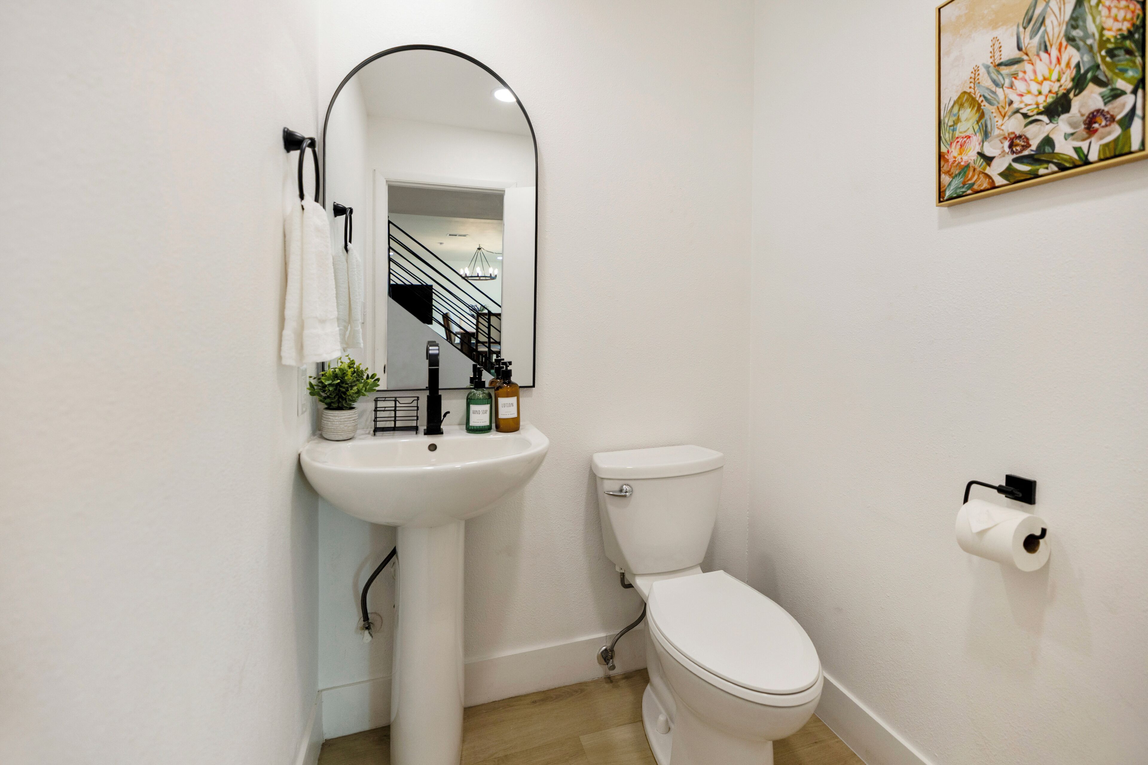 Une salle de bain avec un lavabo blanc, des toilettes et un miroir. On y trouve une plante sur le lavabo, un rouleau de papier toilette accroché au mur et un tableau au-dessus des toilettes.