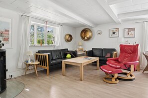 Una sala de estar moderna con un sofá de cuero negro, una mesa de centro de madera y un sillón de cuero rojo. Se ve una ventana con cortinas blancas, una lámpara y una pequeña mesa con un jarrón.