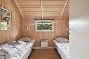 A room with wooden walls, two beds, a window with a view of greenery, and a wall-mounted heater.