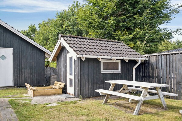 A small wooden shed with a tiled roof and a window, a picnic table, and a bench in front of it.