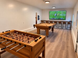 A room with a foosball table, a pool table, and a television displaying a sports game.