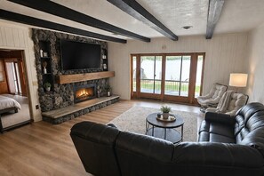 A living room with a stone fireplace, a flat-screen TV, leather sofas, and a large window with a view of the outdoors.