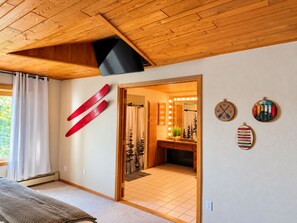 A room with a wooden ceiling, a bed, a mirror with lights, and a ski rack.