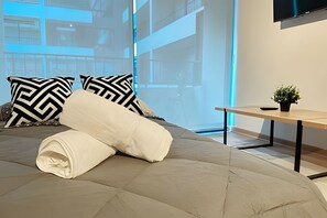 A neatly made bed with a rolled white towel, two black and white patterned pillows, a small potted plant on a wooden side table, and a large window with a view of a building. - Modern apartment near the subway | portable Wi-Fi (Santiago)
