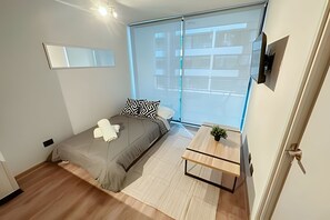 Une chambre à coucher moderne avec un lit, une petite table, une plante, une fenêtre et un miroir.