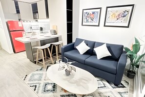 Un salon moderne avec un canapé bleu, une table basse blanche et un coin cuisine à l’arrière-plan.