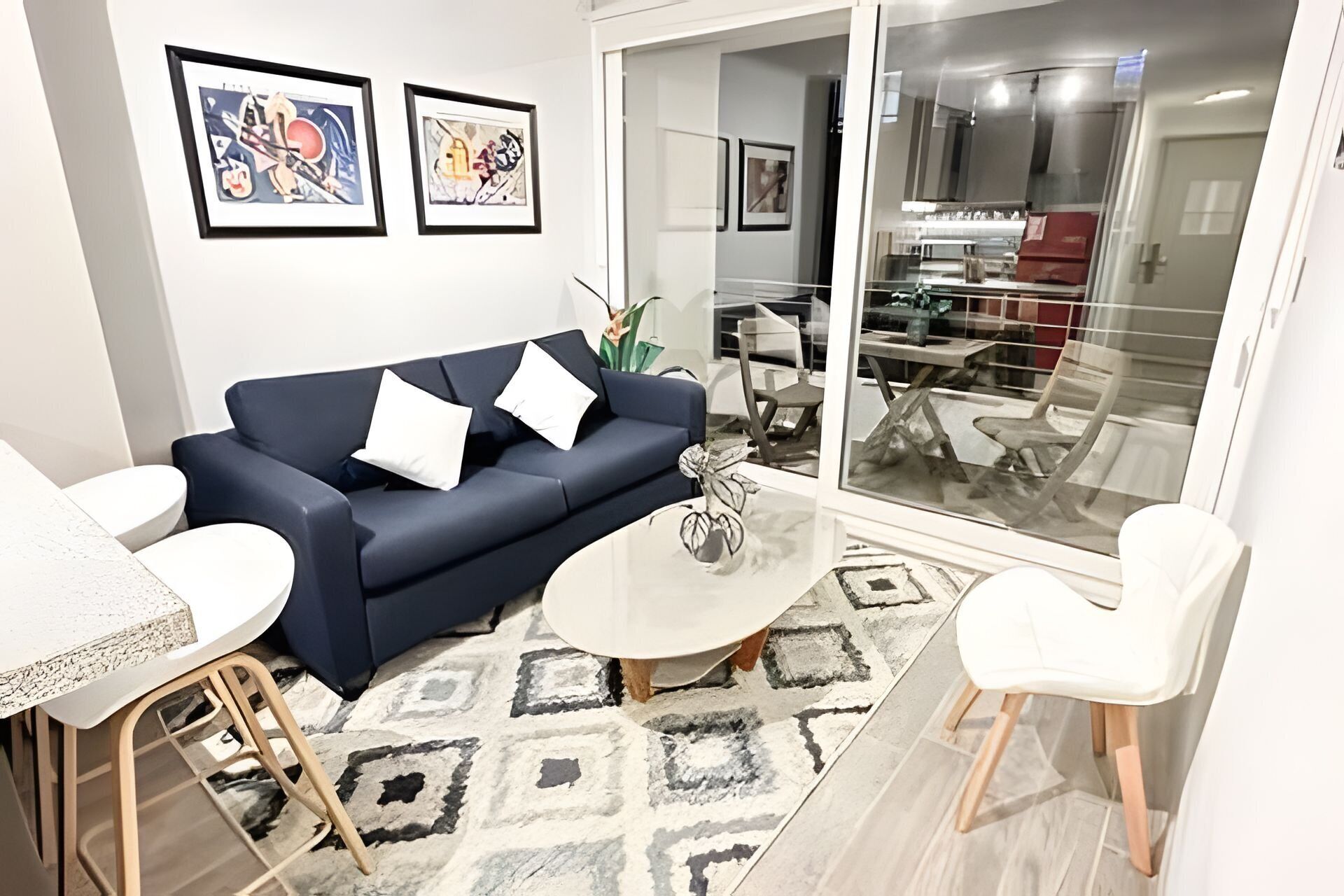 Ein Wohnzimmer mit einem dunkelblauen Sofa, weißen Kissen, einem Couchtisch und einem gemusterten Teppich. An der Wand hängen gerahmte Kunstwerke und eine Glastür führt in einen Essbereich.