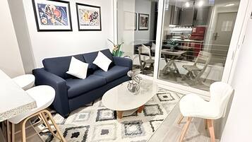 Ein Wohnzimmer mit einem dunkelblauen Sofa, weißen Kissen, einem Couchtisch und einem gemusterten Teppich. An der Wand hängen gerahmte Kunstwerke und eine Glastür führt in einen Essbereich.
