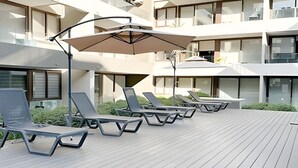 A modern outdoor lounge area with sun loungers and umbrellas, surrounded by a wooden deck and a building with large windows. - Contemporary, comfortable apartment with WiFi and air conditioning (Santiago)