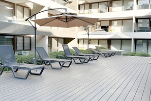 A modern outdoor lounge area with sun loungers and umbrellas, surrounded by a wooden deck and a building with large windows. - Contemporary, comfortable apartment with WiFi and air conditioning (Santiago)