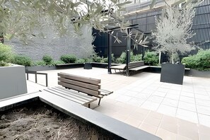 An outdoor seating area with benches and planters, surrounded by greenery and a building with a modern design. - Modern, cozy apartment with WiFi and air conditioning (Santiago)