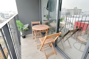 Een balkon met een ronde houten tafel en twee stoelen, een bloempot en uitzicht op gebouwen.