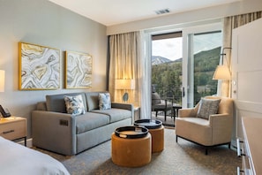 A hotel room with a sofa, armchair, two ottomans, a desk with a lamp, and a view of mountains through a large window. - Gondola Right Outside - Westin Riverfront 2-Bedroom Villa (Avon)