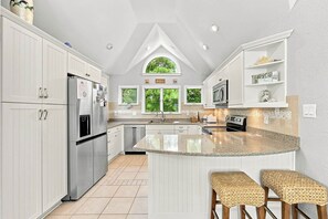 A kitchen with white cabinetry, a stainless steel refrigerator, a granite countertop island, and a window with a view of trees. - Ultra Classic | Private Pool, Hot Tub, and Community Pool (Corolla)