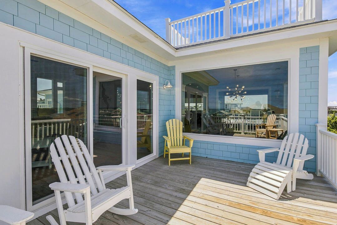 A covered outdoor deck with Adirondack chairs, a glass door entrance, and a view of the interior.