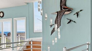 A staircase with metal railings, a wall with a bird sculpture, and a view of the sea through windows. - Sea To Sound | 740 ft to the beach | Private Pool, Hot Tub, and Dog Friendly (Duck)