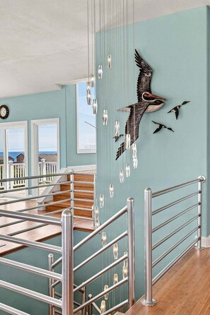 Eine Treppe mit Metallgeländern, eine Wand mit einer Vogel-Skulptur und Blick auf das Meer durch Fenster.