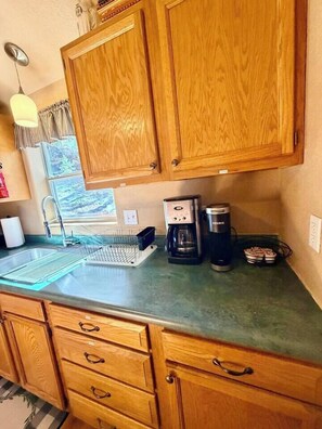 A kitchen with wooden cabinets, a stainless steel sink, a coffee maker, and a window with a view of greenery. - “Peace of the Mountains” (Cloudcroft)