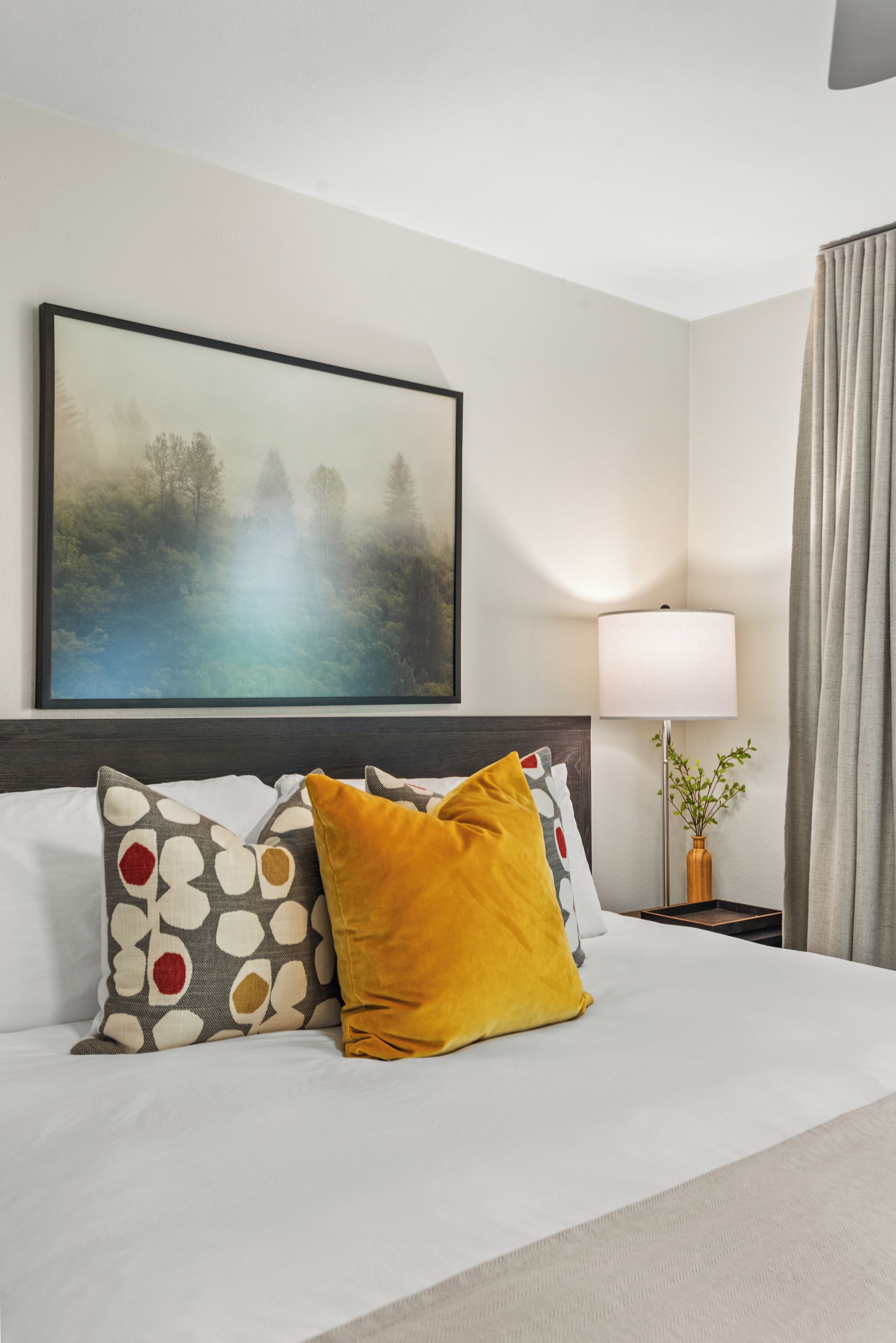 A hotel room with a bed, a painting, a lamp, and a curtain.