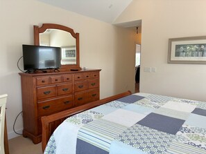 Een slaapkamer met een houten dressoir, een televisie erboven, een grote spiegel en een bed met een geborduurde sprei.