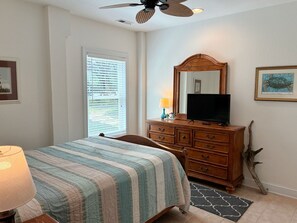 Ein Schlafzimmer mit Bett, Kommode, Fernseher, Deckenventilator, Fenster mit Jalousien und einem gerahmten Bild an der Wand.