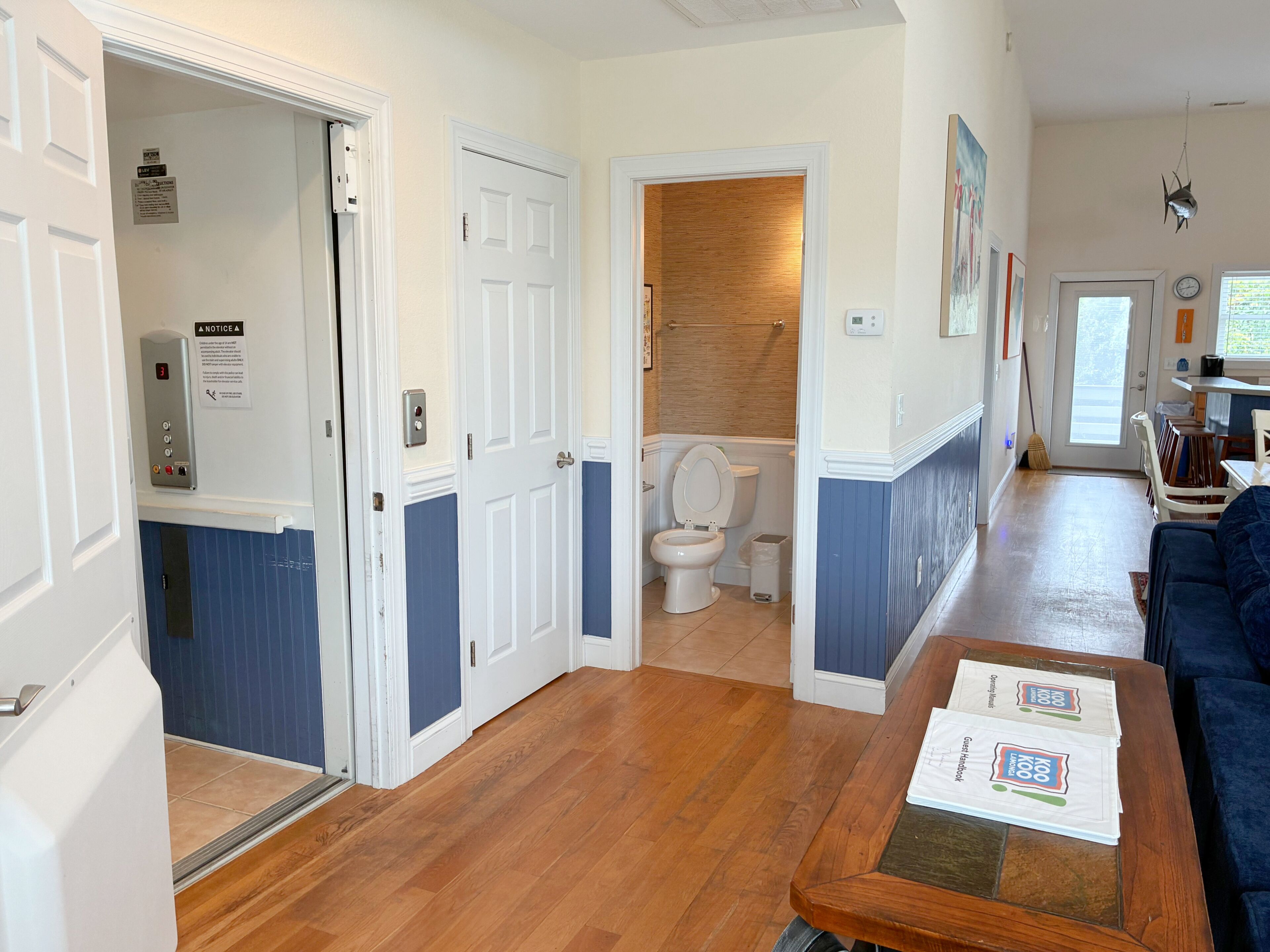 A hotel room with a wooden floor, a white door, a bathroom with a wooden wall, and a blue wall.