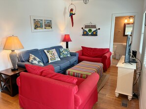 Ein Wohnzimmer mit einem blauen Sofa, roten Sesseln, einem gestreiften Hocker, einer Lampe, einem gerahmten Bild und einer Wanddekoration.