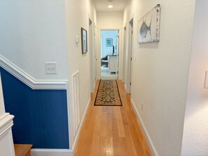 A hallway with a wooden floor, white walls, and a blue accent wall. There is a door at the end of the hallway. - Kookoolamonga!: Oceanside, Pool, Hot Tub, Elevator, Game Room (Nags Head)