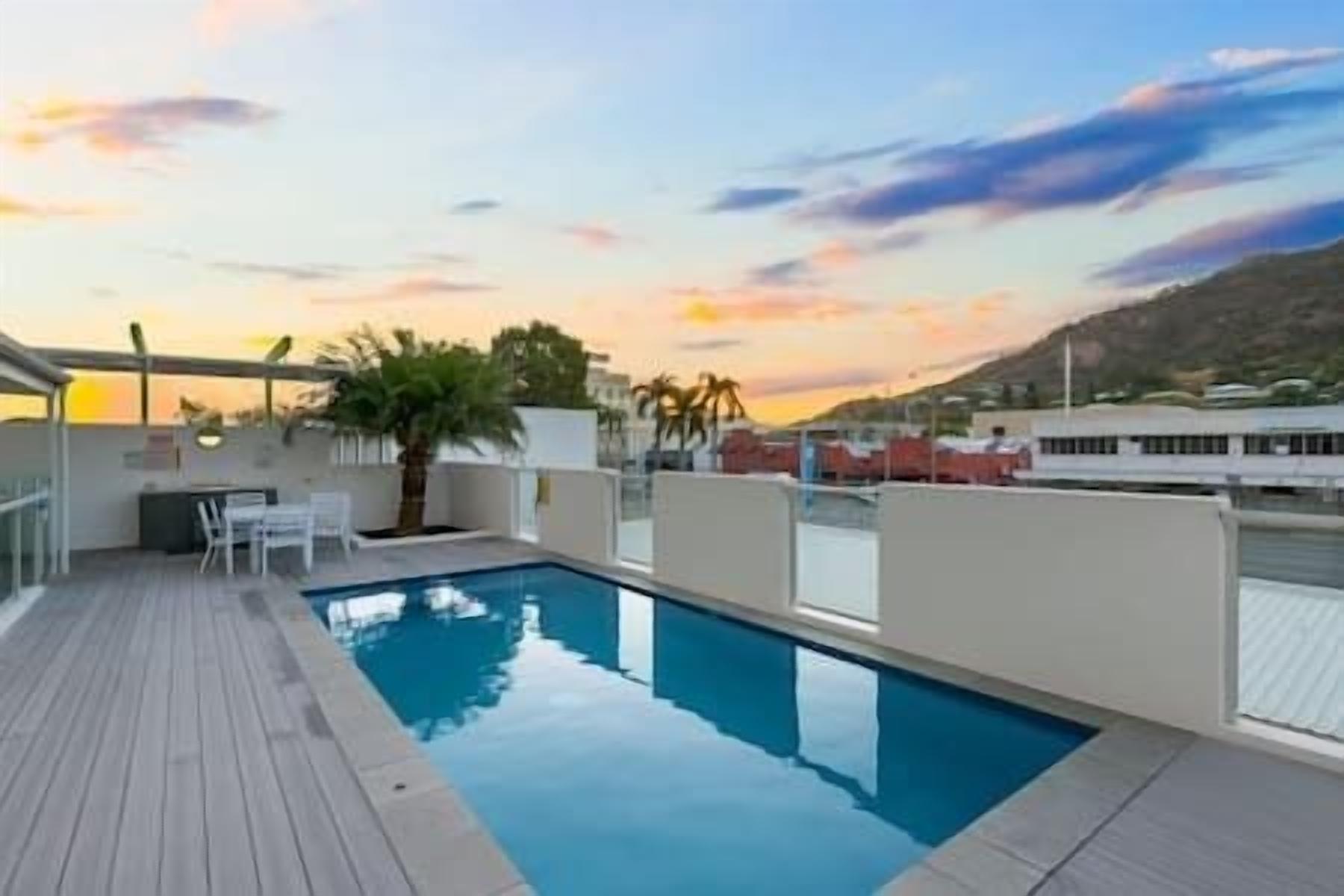 Une piscine sur le toit, dotée d’une terrasse en bois, entourée de murs blancs et surplombant un ciel dégagé.