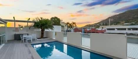 A rooftop pool with a wooden deck, surrounded by white walls and a clear sky.