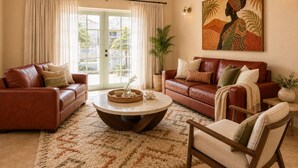 Ein Wohnzimmer mit braunem Ledersofa, einem runden Couchtisch und einem Sessel. An der Wand hängt ein großes Gemälde und ein Fenster mit Vorhängen.