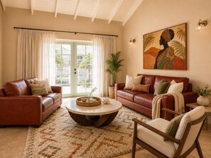 Ein Wohnzimmer mit braunem Ledersofa, einem runden Couchtisch und einem Sessel. An der Wand hängt ein großes Gemälde und ein Fenster mit Vorhängen.