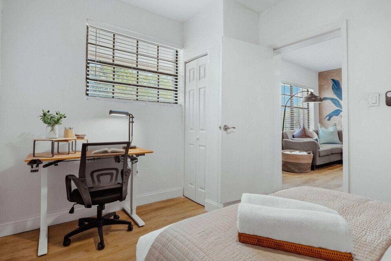 Une chambre à coucher comprenant un lit, un bureau avec une chaise, une fenêtre avec des stores et une porte donnant sur une autre pièce.