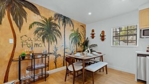 Ein Essbereich mit einer Palmen-Mural und dem Text 'Casa Palma'. Es ist ein Esstisch mit Stühlen, eine Weinsammlung und ein Kühlschrank vorhanden.